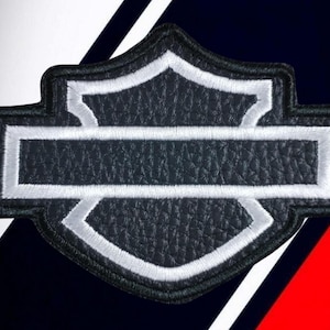 Puede incluir: Un parche bordado en forma de escudo con un centro de piel sintética negra texturizada y un borde cosido en blanco. El diseño consiste en una barra horizontal que cruza una forma de escudo vertical, todo delineado con costuras negras.
