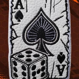 May include: A rectangular embroidered patch featuring a white faux leather background with a black ace of spades playing card design. The patch displays a large central spade symbol, the letter A in the corners, and a six-sided die in the foreground.