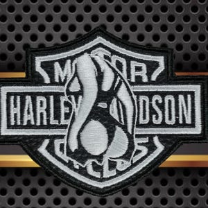May include: Embroidered Harley-Davidson patch with the classic logo in black and white. The patch is shield-shaped, displaying the words "HARLEY-DAVIDSON" and "MOTOR". Set against a textured, dark grey background with a gold stripe.