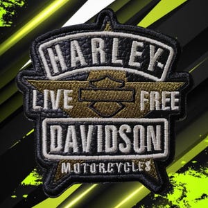 Może przedstawiać: Czarno-złota haftowana naszywka z napisem HARLEY DAVIDSON, LIVE FREE oraz MOTORCYCLES. Wzór zawiera centralny emblemat tarczy ze skrzydłami, białe litery na czarnym fakturowanym tle oraz akcenty wykonane złotą nicią metaliczną.
