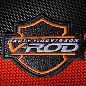 May include: A black leather-textured patch shaped like a shield with orange embroidered borders. The text HARLEY-DAVIDSON is stitched in white above the large, stylized silver and orange V-ROD lettering, all centred within the shield design.