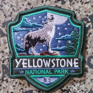 May include: A shield-shaped embroidered patch featuring a gray wolf howling at a large white moon against a dark blue starry night sky with green pine trees. The text reads YELLOWSTONE NATIONAL PARK in white letters on a dark green background.