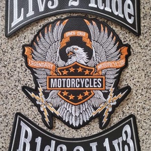 Peut inclure: Un ensemble de trois écussons brodés noirs avec des détails blancs et orange. Le rocker supérieur indique L1v3 2 Ride, le rocker inférieur indique R1d3 2 L1v3, et l'écusson central présente un aigle, des éclairs et le texte Legendary Motorcycles.