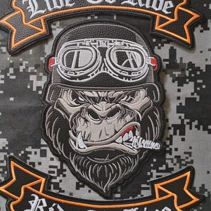 Puede incluir: Un parche bordado negro que muestra un gorila con casco y gafas, con un cigarro en la boca. La cinta superior dice "Live To Ride" y la cinta inferior dice "Ride To Live" en letras blancas con bordes de color naranja.