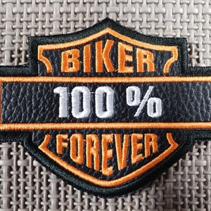 Puede incluir: Un parche de motero negro y naranja con las palabras "BIKER 100% FOREVER" en letras blancas y naranjas. El parche tiene forma de escudo con borde negro y fondo texturizado.