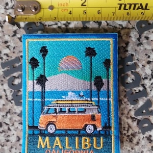 Può includere: Una toppa ricamata rettangolare raffigurante un furgoncino arancione vintage con una tavola da surf sul tetto, palme, un tramonto e la scritta MALIBU CALIFORNIA. Un metro a nastro giallo indica un'altezza di circa 8,9 cm.