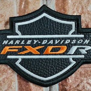 May include: A black shield-shaped embroidered patch with a textured faux leather appearance. It features the white text HARLEY-DAVIDSON above the orange and silver text FXDR, all enclosed within a white and black stitched border design.