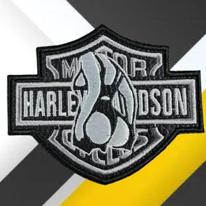 Könnte beinhalten: Schwarzer und grauer Harley-Davidson-Aufnäher mit Schilddesign. Der Aufnäher zeigt das Harley-Davidson-Logo in weißer Schrift und ein stilisiertes Bild eines Motorradmotors. Der Aufnäher ist schwarz umrandet.