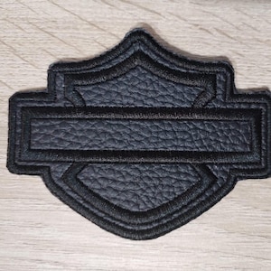 May include: A black shield-shaped patch featuring a textured faux leather surface with a horizontal bar across the center. The entire perimeter and the central bar are outlined with thick, raised black embroidery stitching, creating a defined border.