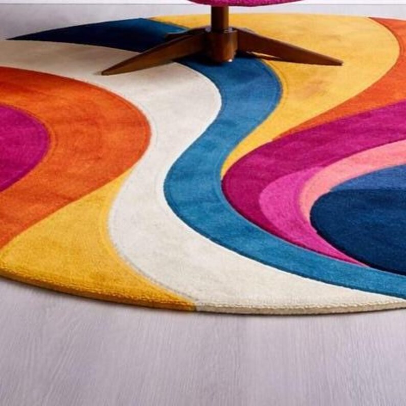 May include: A round, colorful rug with a retro design. The rug features a wavy pattern in shades of orange, yellow, pink, blue, and white. A wooden chair leg is visible at the top. The rug is a vibrant addition to any room.