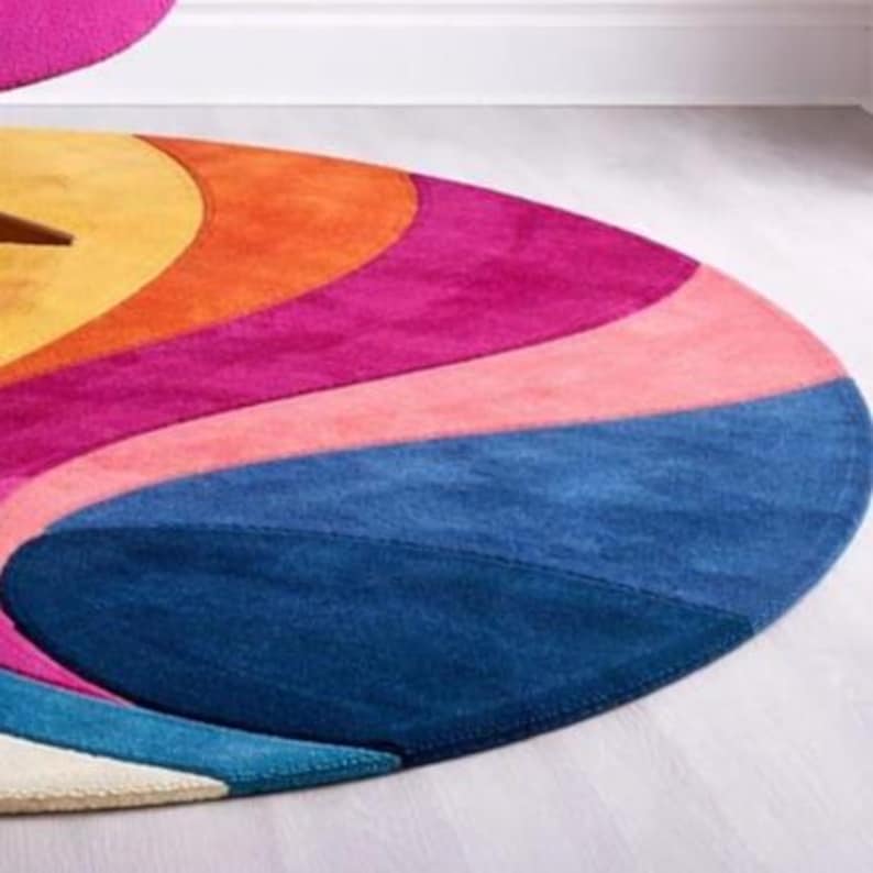 May include: A colorful, oval-shaped rug with a vibrant design. The rug features a spectrum of colors, including yellow, orange, pink, purple, and blue. The rug is made of a soft material and is likely used for home decor.