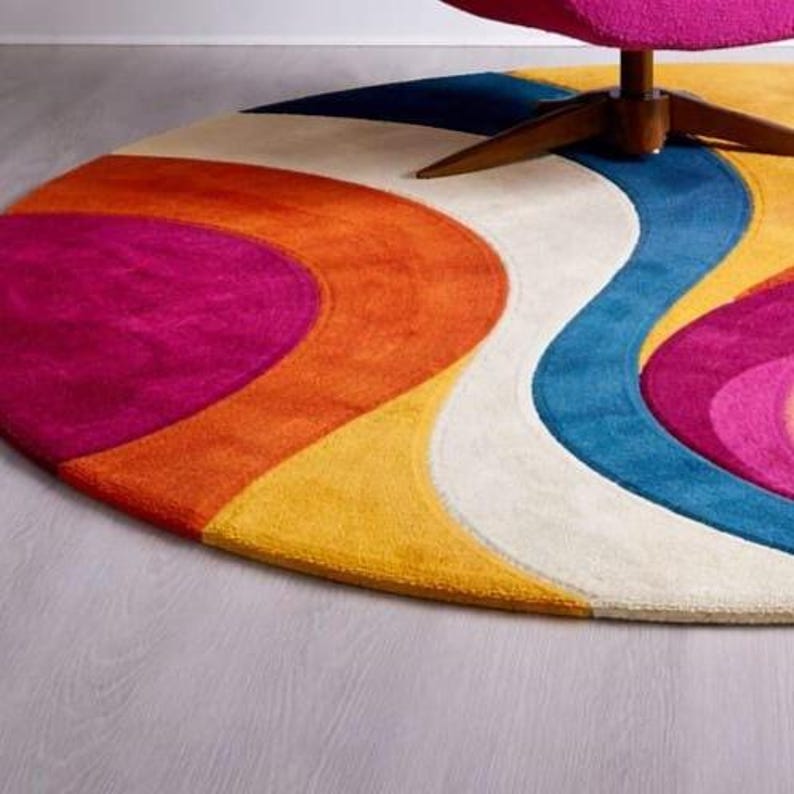 May include: A colorful, round area rug with a retro design. The rug features a vibrant pattern of curved stripes in shades of pink, orange, yellow, blue, and white. The rug is placed on a light gray floor, with a portion of a chair visible.