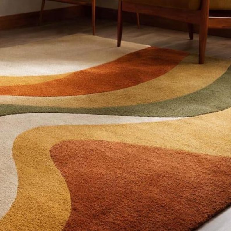 May include: A retro-style area rug with a wavy design in shades of cream, yellow, orange, and green. The rug has a soft, textured appearance and is placed on a wooden floor. The design evokes a vintage aesthetic.
