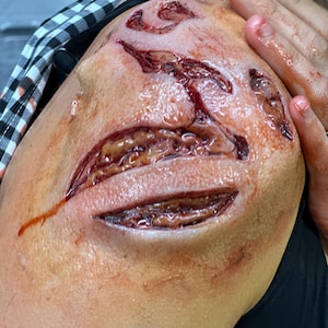 May include: A close-up of a shoulder with realistic special effects makeup depicting deep, open wounds. The wounds are red and appear fresh, with exposed tissue and blood. The skin around the wounds is a pale flesh tone.