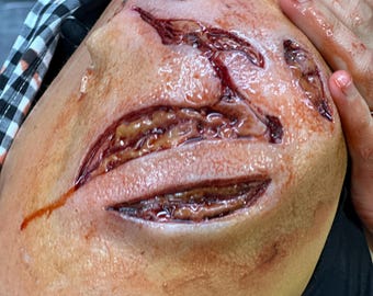 May include: A close-up of a shoulder with realistic special effects makeup depicting deep, open wounds. The wounds are red and appear fresh, with exposed tissue and blood. The skin around the wounds is a pale flesh tone.