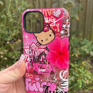 May include: Pink phone case with a vibrant design featuring a Hello Kitty character, a large pink hibiscus flower, and the word "Juicy" in a cursive font. The case has a glossy finish and a black camera cutout.