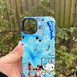 K&ouml;nnte beinhalten: Eine blaue Handyh&uuml;lle mit floralem Design, Schmetterlingen, Sternen und einer Hello Kitty-Grafik. Die H&uuml;lle hat einen schwarzen Rand. Das Design beinhaltet Blau- und Wei&szlig;t&ouml;ne. Die H&uuml;lle wird in einer Hand gehalten.