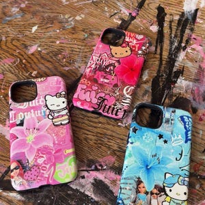 May include: Three colorful phone cases with unique designs. One is pink with a Hello Kitty graphic and floral accents, another is pink with the word "Juicy", and the third is blue with a floral design and a Hello Kitty graphic. The cases are on a wooden surface.
