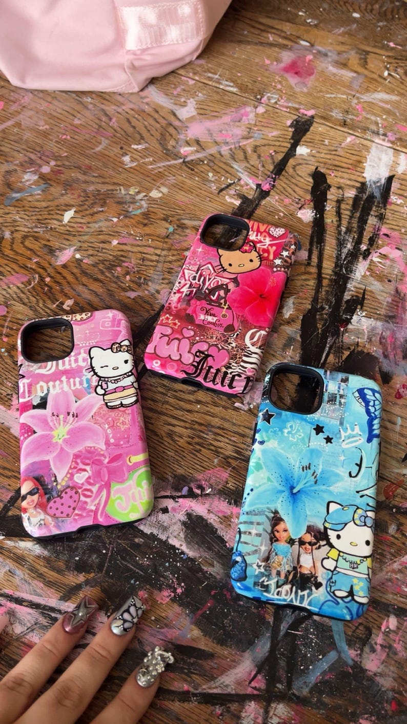 May include: Three colorful phone cases with unique designs. One is pink with a Hello Kitty graphic and floral accents, another is pink with the word "Juicy", and the third is blue with a floral design and a Hello Kitty graphic. The cases are on a wooden surface.