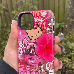May include: A pink phone case featuring a Hello Kitty graphic, a large pink hibiscus flower, and the words "Juicy Couture". The case has a glossy finish and a black camera cutout.