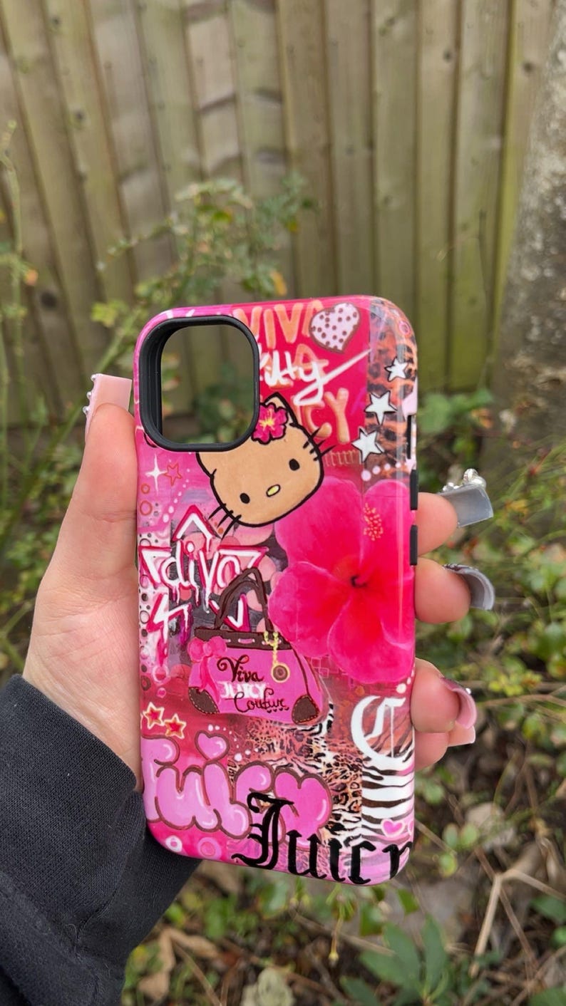 May include: A pink phone case featuring a Hello Kitty graphic, a large pink hibiscus flower, and the words "Juicy Couture". The case has a glossy finish and a black camera cutout.