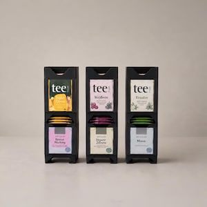 Könnte beinhalten: Drei schwarze vertikale Teebeutelspender, die jeweils zwei Stapel Teebeutel enthalten. Die Etiketten zeigen den Markennamen tee freude und verschiedene Geschmacksrichtungen wie Beeren Mischung, Ingwer Zitrone, Waldbeere, Kräuter und Minze mit Abbildungen.