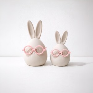 Könnte beinhalten: Zwei Keramik-Hasenfiguren. Die Hasen sind eiförmig mit langen Ohren und tragen rosafarbene Brillen. Die Hasen sind hellbeige und stehen vor einem weißen Hintergrund.