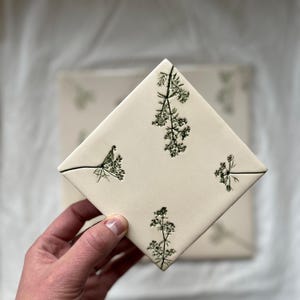 Op de afbeelding: Een vierkante, crèmekleurige keramische tegel met een botanisch ontwerp van groene takjes. De tegel wordt in een hand gehouden, met een andere tegel zichtbaar op de achtergrond. De takjes zijn gerangschikt in een herhalend patroon over het oppervlak van de tegel.