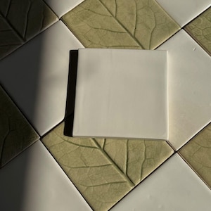 Puede incluir: Un azulejo de cerámica cuadrado y liso de color blanco roto colocado en el centro sobre una cuadrícula de azulejos alternos en blanco roto y verde salvia claro, cada uno con un patrón detallado de nervaduras de hoja en relieve en diagonal.