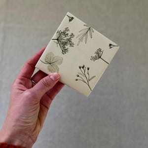 Könnte beinhalten: Eine quadratische, cremefarbene Keramikfliese mit einem botanischen Design aus Blättern und Zweigen in Grün und Grau. Die Fliese wird in einer Hand vor einem neutralen Hintergrund gehalten und zeigt das detaillierte Blumenmuster.
