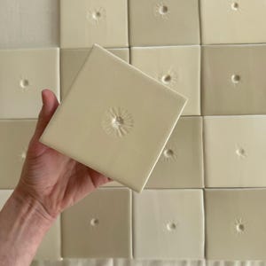 Może przedstawiać: Kolekcja kwadratowych, złamanych białych płytek ceramicznych ułożonych w siatkę. Każda płytka posiada mały, centralnie umieszczony wytłoczony detal w kształcie promieni słońca. Płytki mają gładkie, błyszczące wykończenie i są jednolite pod względem kształtu.
