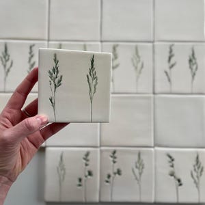 Puede incluir: Azulejos cuadrados de cerámica blanca con un diseño botánico dibujado a mano. Cada azulejo presenta una delicada ilustración verde y gris de una planta. Los azulejos están dispuestos en un patrón de cuadrícula, con un azulejo en la mano.