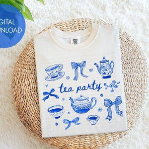 May include: Cream-colored t-shirt with a blue tea party design. The design includes teacups, teapots, bows, and the words "tea party". The shirt is folded on a woven mat.