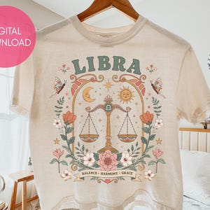 May include: A beige t-shirt featuring a graphic print with the word LIBRA in green letters. The design includes a central scale, a sun, a moon, butterflies, and floral elements in shades of orange, pink, and green, with the text BALANCE HARMONY GRACE.