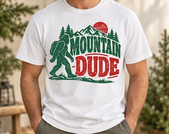 Puede incluir: Una camiseta blanca de manga corta con un estampado gráfico de una silueta verde de Bigfoot caminando frente a una cordillera con pinos y un sol rojo. El texto "MOUNTAIN DUDE" está impreso en letras grandes y desgastadas de color rojo y verde.