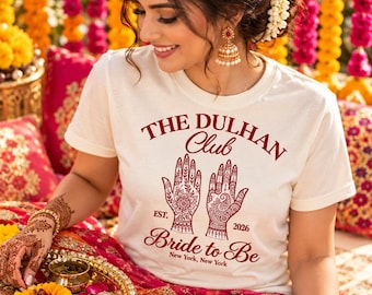 Puede incluir: Una camiseta de manga corta color crema con texto en rojo oscuro que dice THE DULHAN Club, EST. 2026, Bride to Be y New York, New York. El gráfico central muestra dos manos decoradas con intrincados patrones de henna en el pecho.