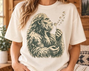 Puede incluir: Una camiseta de manga corta color crema con una ilustración gráfica en verde oscuro de una criatura peluda que se asemeja a un Bigfoot soplando un diente de león, con semillas flotando desde la flor en una curva hacia la derecha.
