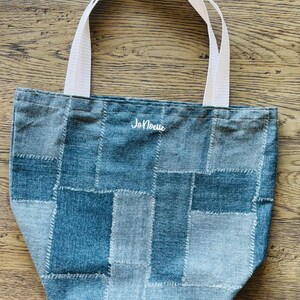 May include: A denim tote bag with a patchwork design in shades of blue and gray. The bag features white handles and the name "JoNoelle" embroidered in white thread. The bag is designed for carrying items.