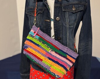 Könnte beinhalten: Eine bunte, rechteckige Häkeltasche mit langem Riemen. Die Tasche hat eine lebendige Mischung aus Regenbogenfarben, darunter Lila, Grün, Gelb, Orange und Rosa. Die Tasche wird auf einer Schaufensterpuppe mit Jeansjacke und rotem Rock präsentiert.