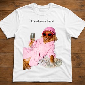 Op de afbeelding: Een wit t-shirt met een afbeelding van een hond in een roze badjas, een roze baseballpet en een rode zonnebril. De hond houdt een glas champagne vast en rust met zijn poot op een waaier van bankbiljetten. De tekst "I do whatever I want" staat erboven.
