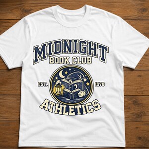 Könnte beinhalten: Ein weißes Kurzarm-T-Shirt mit einem Grafikdesign, das den Text MIDNIGHT BOOK CLUB ATHLETICS und EST. 1978 zeigt. Die zentrale kreisförmige Illustration stellt ein offenes Buch, eine leuchtende Laterne, eine Mondsichel und Sterne dar.