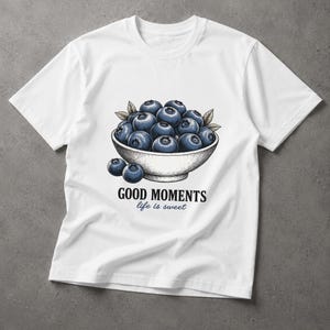 May include: A white short-sleeve t-shirt featuring a graphic illustration of a bowl filled with dark blue blueberries. Below the bowl, the text reads GOOD MOMENTS in bold black letters and life is sweet in a smaller blue script font.