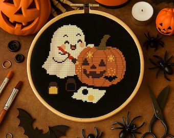 May include: A circular wooden embroidery hoop containing a black fabric piece with a cross-stitch design of a white ghost carving an orange jack-o'-lantern. The design includes small paint pots and a palette, surrounded by Halloween-themed craft supplies.