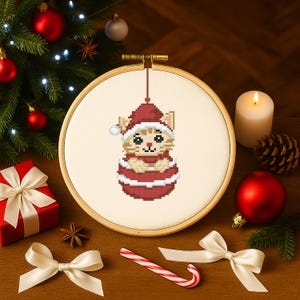 May include: A cross-stitch Christmas ornament featuring a cat wearing a Santa hat and suit, displayed in a wooden embroidery hoop. The ornament is surrounded by festive decorations, including a lit candle, a gift box, and a candy cane.