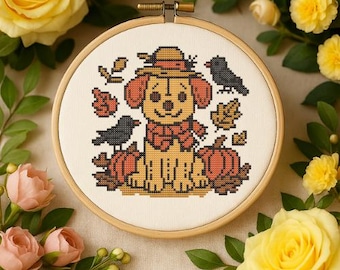 May include: A circular wooden embroidery hoop containing a cross-stitch pattern of a tan puppy wearing a hat and a red scarf, surrounded by two black birds, orange pumpkins, and scattered autumn leaves on a white fabric background.