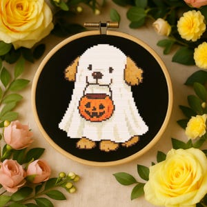 May include: A cross-stitch embroidery piece in a round wooden hoop featuring a dog wearing a white ghost costume and holding an orange jack-o'-lantern candy bucket. The design is stitched on a solid black fabric background with visible stitches.