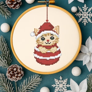 Può includere: Ricamo a punto croce di un gatto con cappello e abito da Babbo Natale, presentato in un telaio di legno. Il gatto è beige e rosso, con occhi verdi. Lo sfondo è verde acqua, decorato con aghi di pino, pigne e ornamenti.