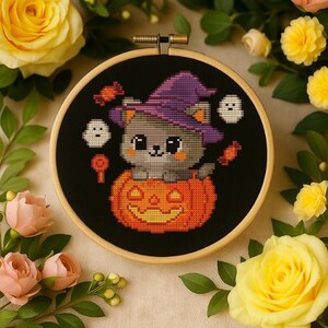 May include: A cross-stitch embroidery in a wooden hoop shows a grey cat in a purple witch hat, inside an orange pumpkin. Two white ghosts and sweets surround the cat on a black background. The embroidery is surrounded by yellow and pink roses.