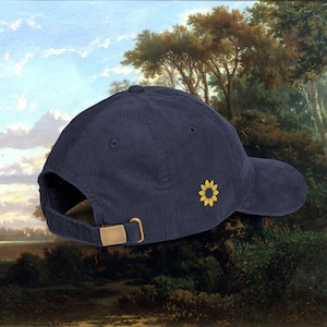 Puede incluir: Gorra de béisbol de pana azul marino con visera curva y hebilla ajustable dorada. Una pequeña flor de girasol amarilla está bordada en el lateral. La gorra está sobre un cuadro de paisaje.