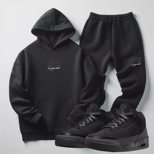 Puede incluir: Conjunto negro de sudadera con capucha y pantalón de chándal, ambos con el texto "23 Legend" en blanco. La sudadera tiene un bolsillo delantero. Zapatillas deportivas negras de caña alta completan el conjunto.